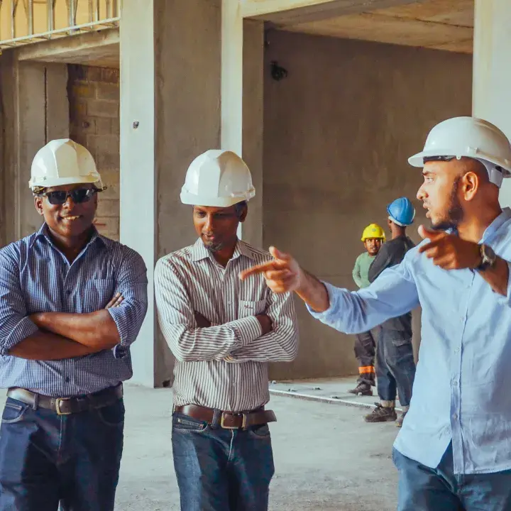 Three men in white hard hats discuss a concrete building under construction as one points, with workers in the background