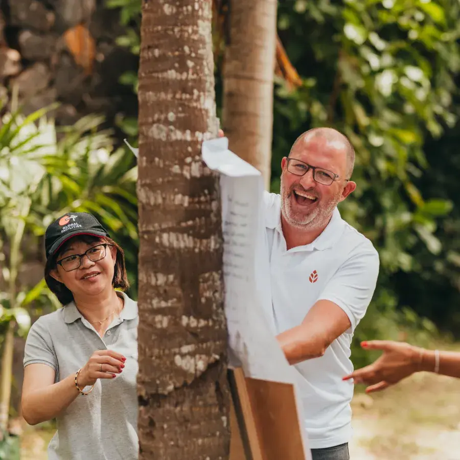 Laughing Novaterra team members pin a paper sheet to a palm tree during an outdoor team-building activity