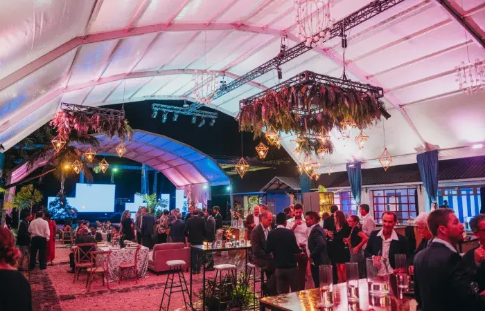 Guests mingling at an evening launch event under a lit marquee with hanging floral chandeliers and a stage screen, 2019