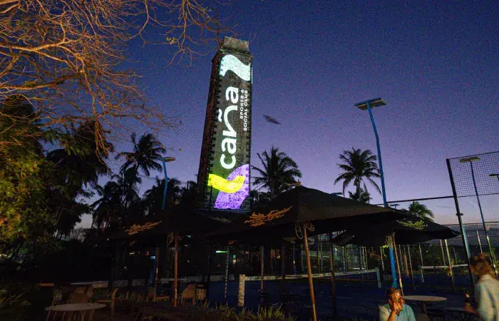 Old sugar mill chimney lit at night with a Caña Sports and Social Club projection, above parasols and padel courts