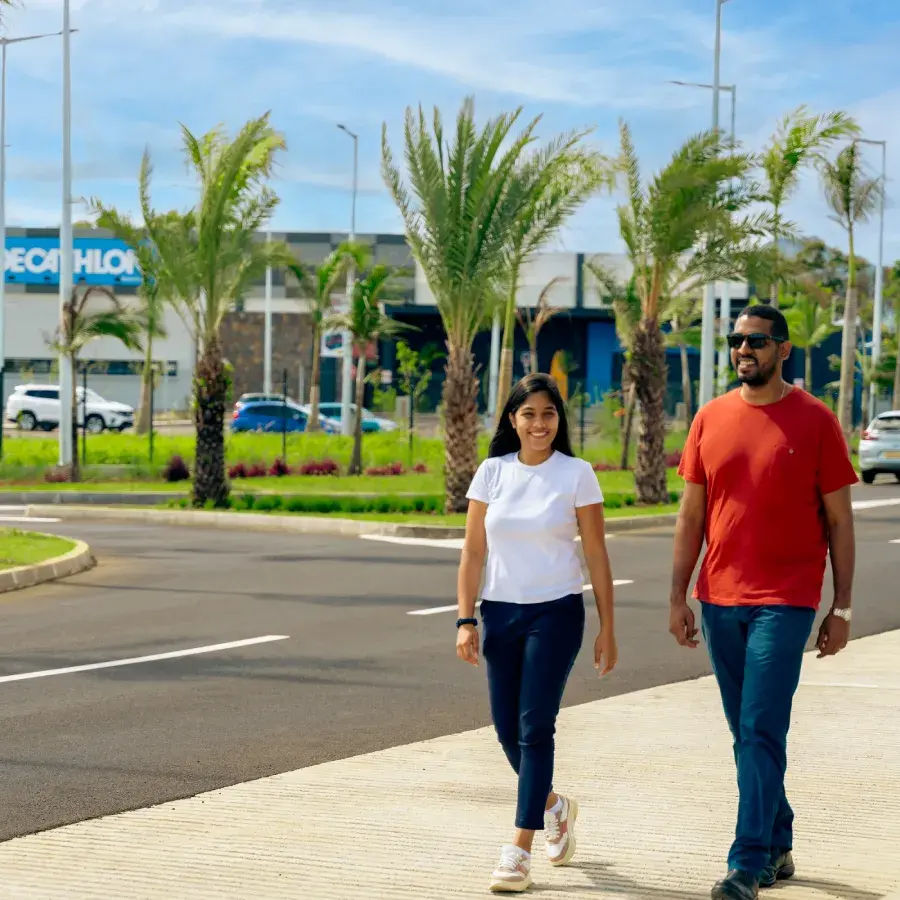Un couple se promène sur un trottoir bordé de palmiers du Business District de Beau Plan, devant un magasin Decathlon