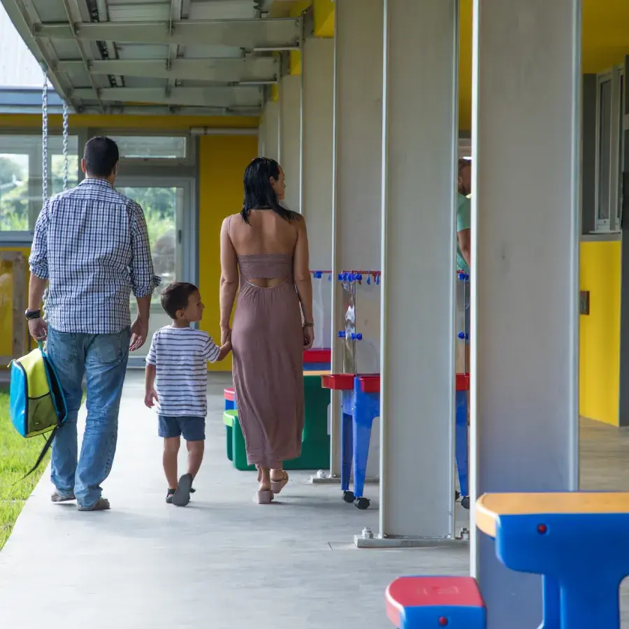 Un couple et leur jeune fils marchent sous la galerie couverte d'une école colorée du quartier éducatif de Beau Plan