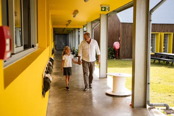 Un homme et une fillette en uniforme marchent main dans la main sous la galerie jaune d’une école de Beau Plan