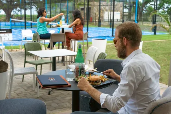 Un homme déjeune en terrasse face aux courts de padel de Beau Plan, où deux femmes discutent à une table voisine.