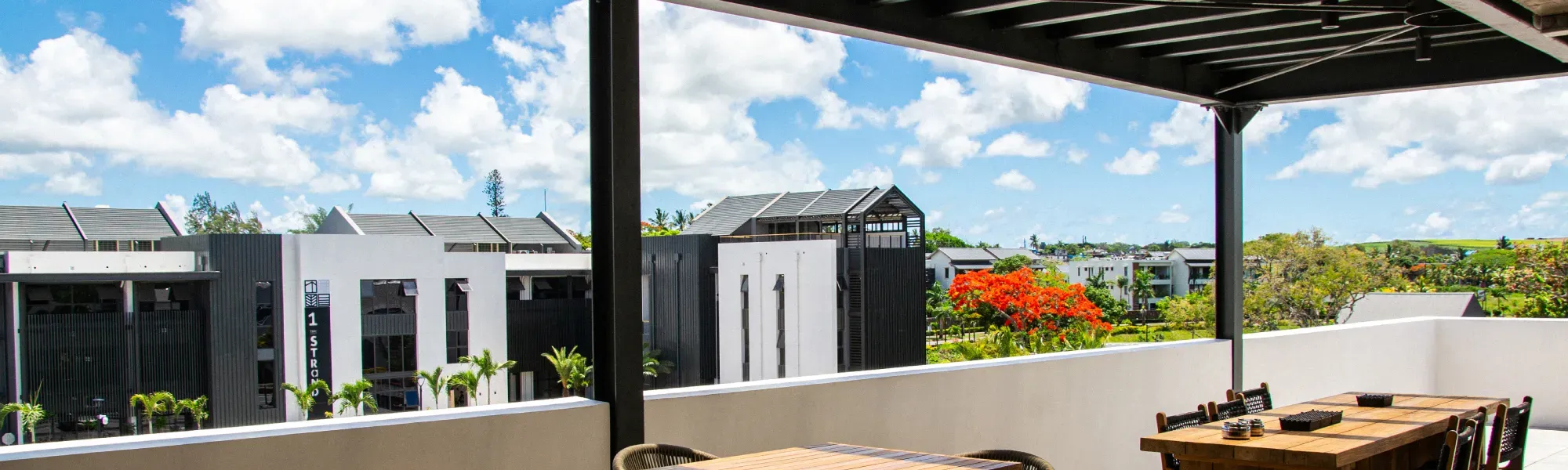 Terrasse couverte avec tables en bois donnant sur des immeubles résidentiels modernes et un flamboyant en fleurs à Maurice