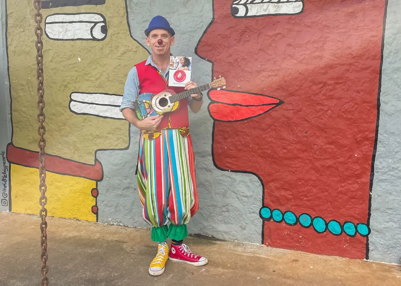 Mediclown clown with red nose, striped trousers and ukulele holding a leaflet in front of a colourful mural of faces