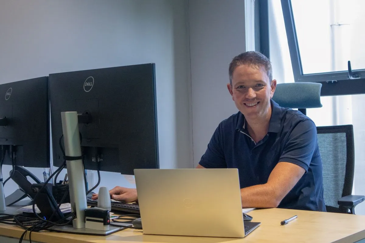 Will Morgan of Mourant Global Services smiling at his desk with a laptop and Dell monitors in The Strand offices, Beau Plan