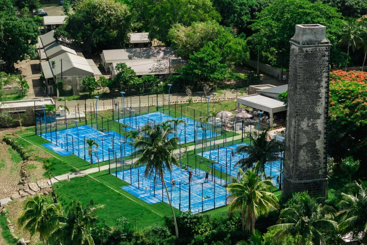 Aerial view of Isla Padel's blue padel courts in Beau Plan beside an old stone sugar mill chimney and palm trees