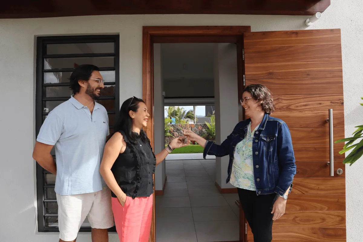 A woman hands house keys to a smiling couple at the wooden front door of a new Mango Village Phase 2 home in Beau Plan