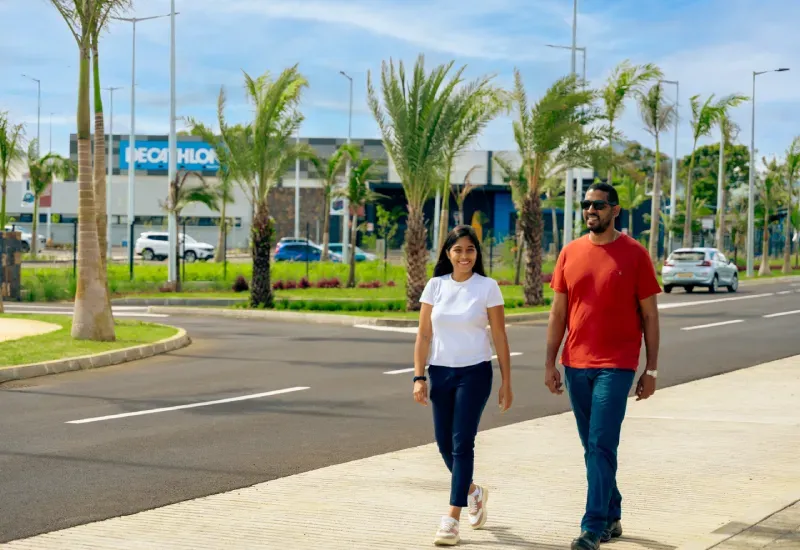 Two people walking along a palm-lined avenue in Beau Plan Business District, with a Decathlon store in the background
