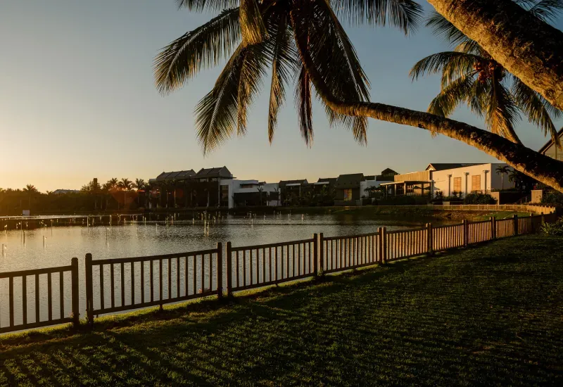Mahogany Shopping Promenade buildings across the Beau Plan lake at sunset, framed by palm trees and a wooden fence