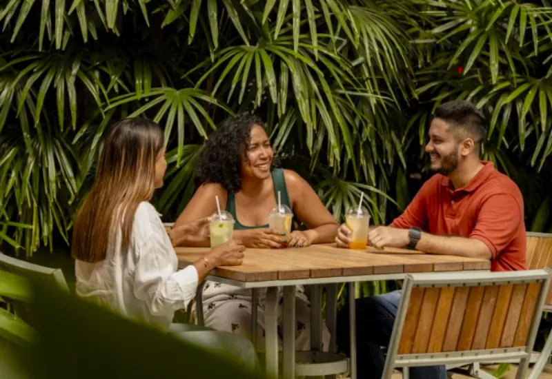 Three friends laugh over iced drinks at a wooden terrace table among palms at Mahogany Shopping Promenade