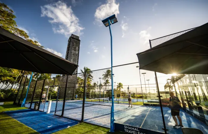 Players on the outdoor glass-walled courts of Caña Padel Club at Mon Rocher, with the historic sugar estate chimney behind
