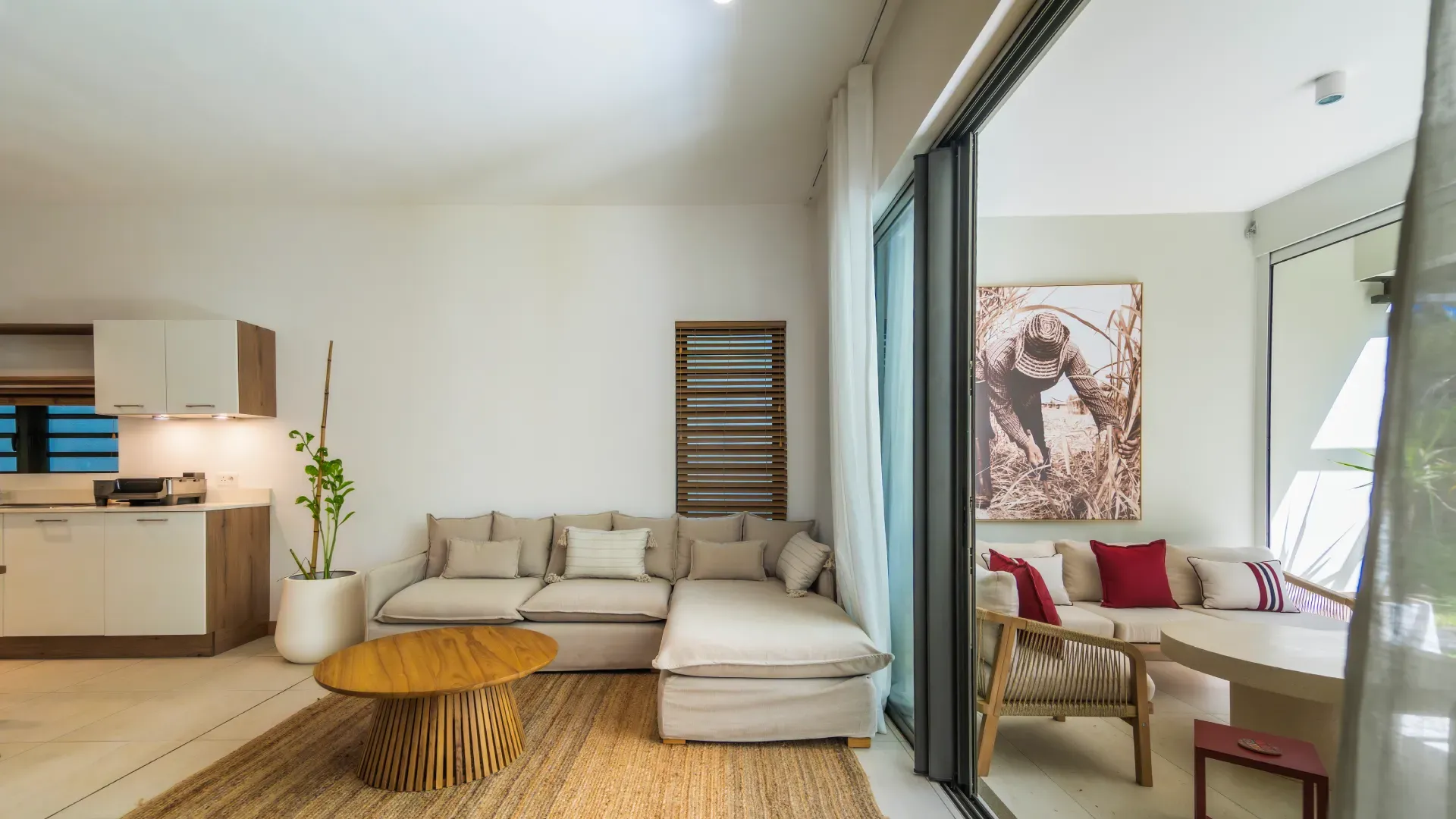 Living room of a 3-bedroom Mango Village apartment with beige sofa, open kitchen and terrace with red cushions and art