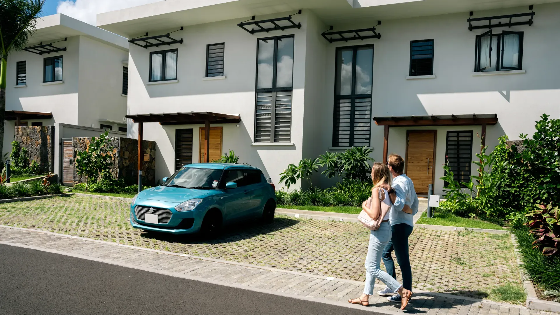 Couple admiring white two-storey duplex homes with wooden pergolas and a blue car parked in Mango Village, Beau Plan