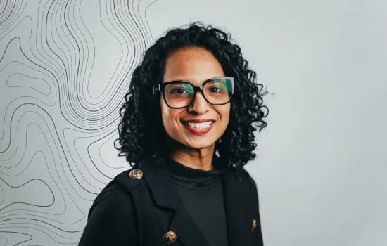 Portrait of Jovanah Poonoosamy, Novaterra accountant, smiling in glasses and a black jacket before a contour-line wall