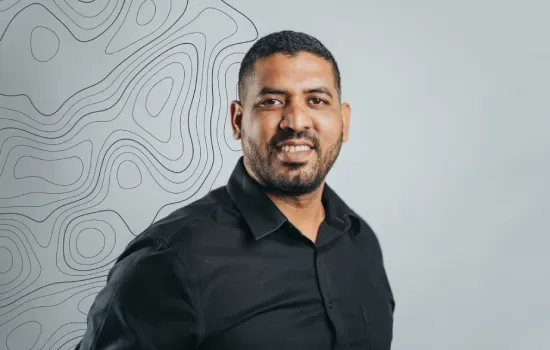 Portrait of Evam Bissoon, Health & Safety Coordinator, smiling in a black shirt against a grey topographic-line backdrop
